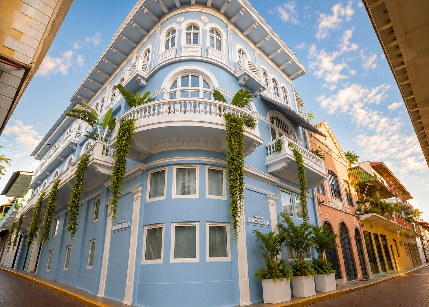 Edificio azul en esquina con detalles blancos, balcones y hiedra en una calle soleada bajo un cielo azul.