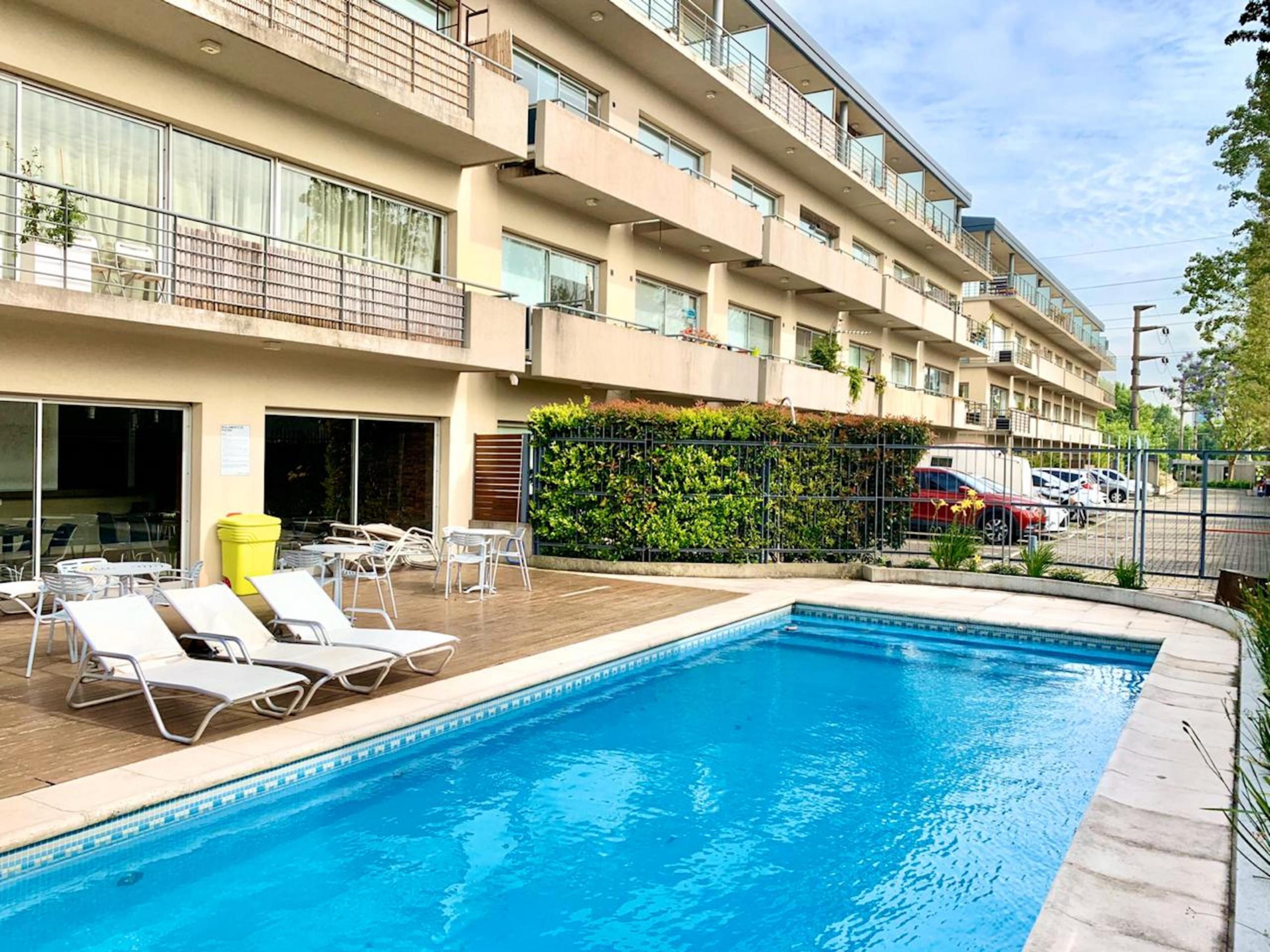 A pool in front of a three-story beige building with balconies, patio furniture, and a green wall.