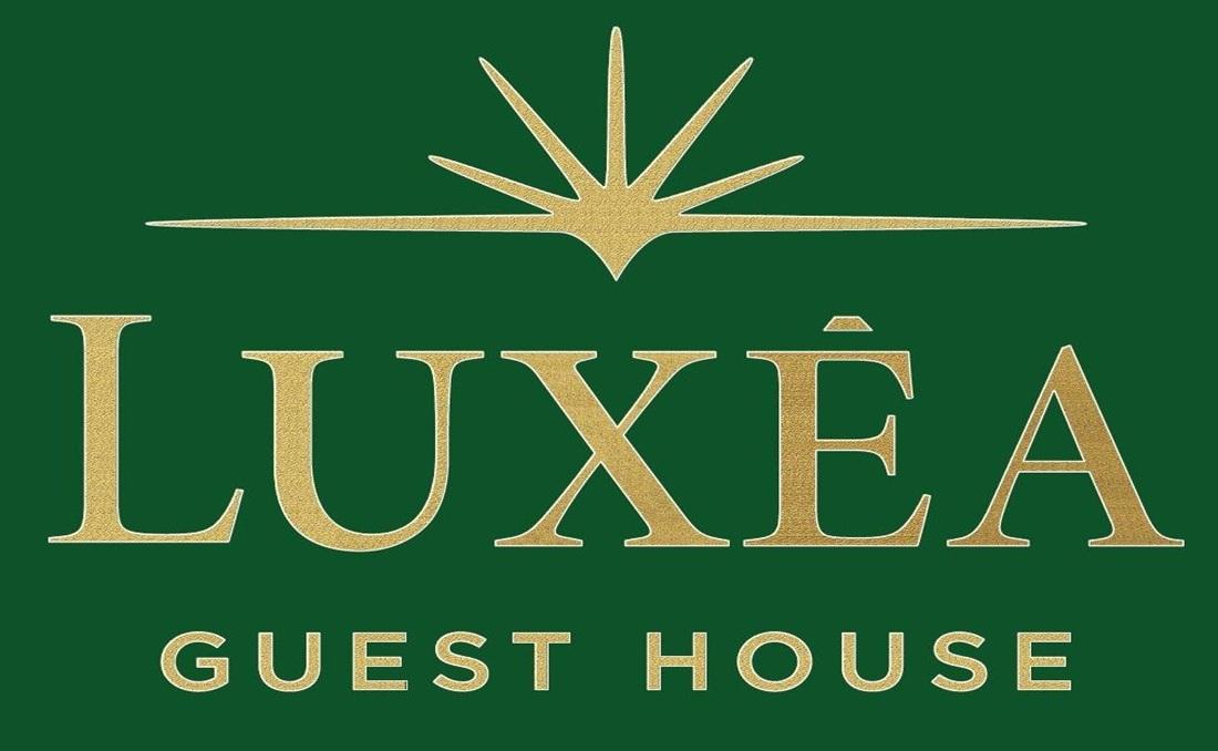 Logo per Lux&eacute;a Guest House con testo serif dorato e un'icona a forma di raggio di sole su uno sfondo rettangolare verde scuro